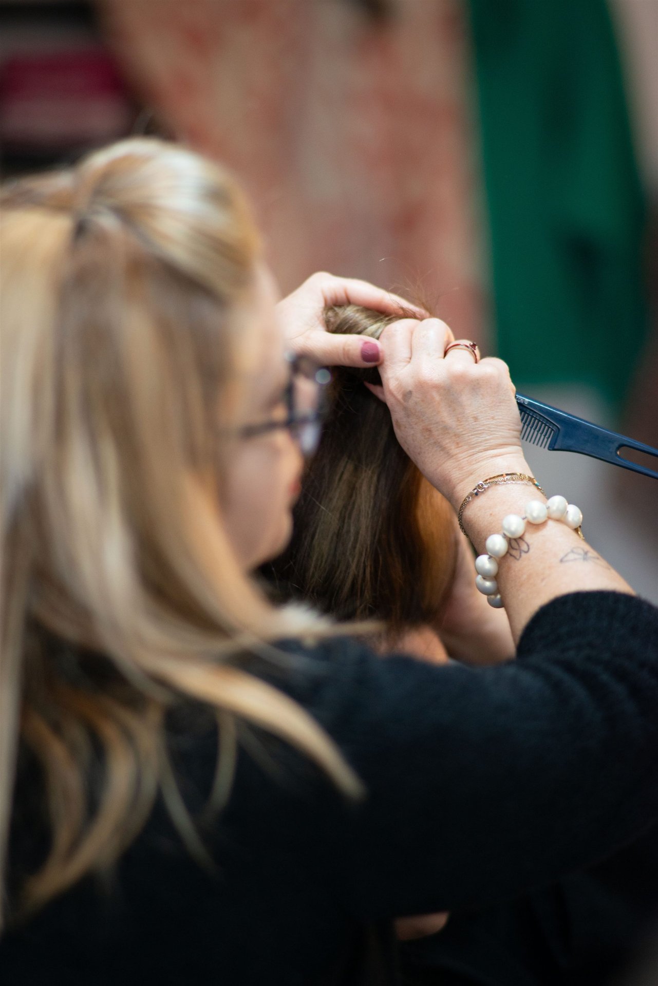 Annabel de dos travaille le chignon d'Amandine avec un peigne bleu — bracelets perles et argent visibles