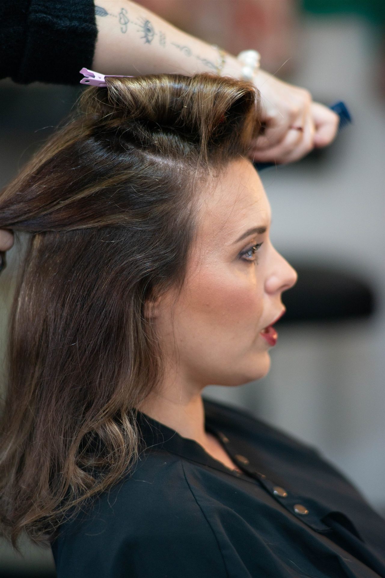 Profil d'Amandine pendant la coiffure — mains d'Annabel travaillent le chignon, regard à droite