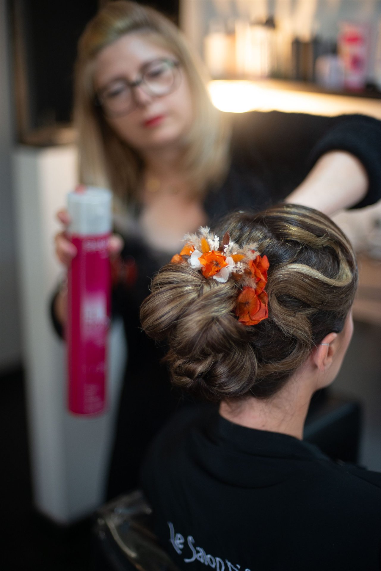 Annabel pose du laque sur le chignon fleuri d'Amandine — fleurs oranges et blanches de Mille Fleurs Sauvages