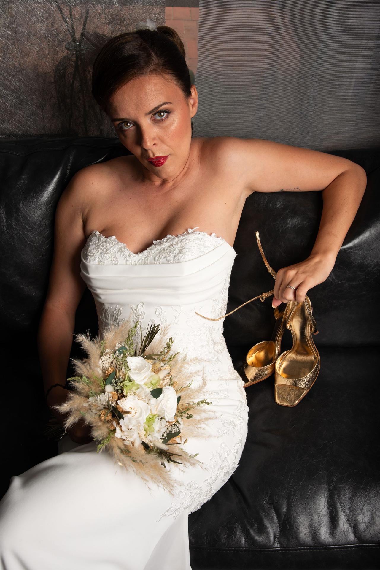 Portrait de la mariée allongée sur canapé noir, bouquet blanc pampas et talons dorés à la main — shooting mariage Perpignan