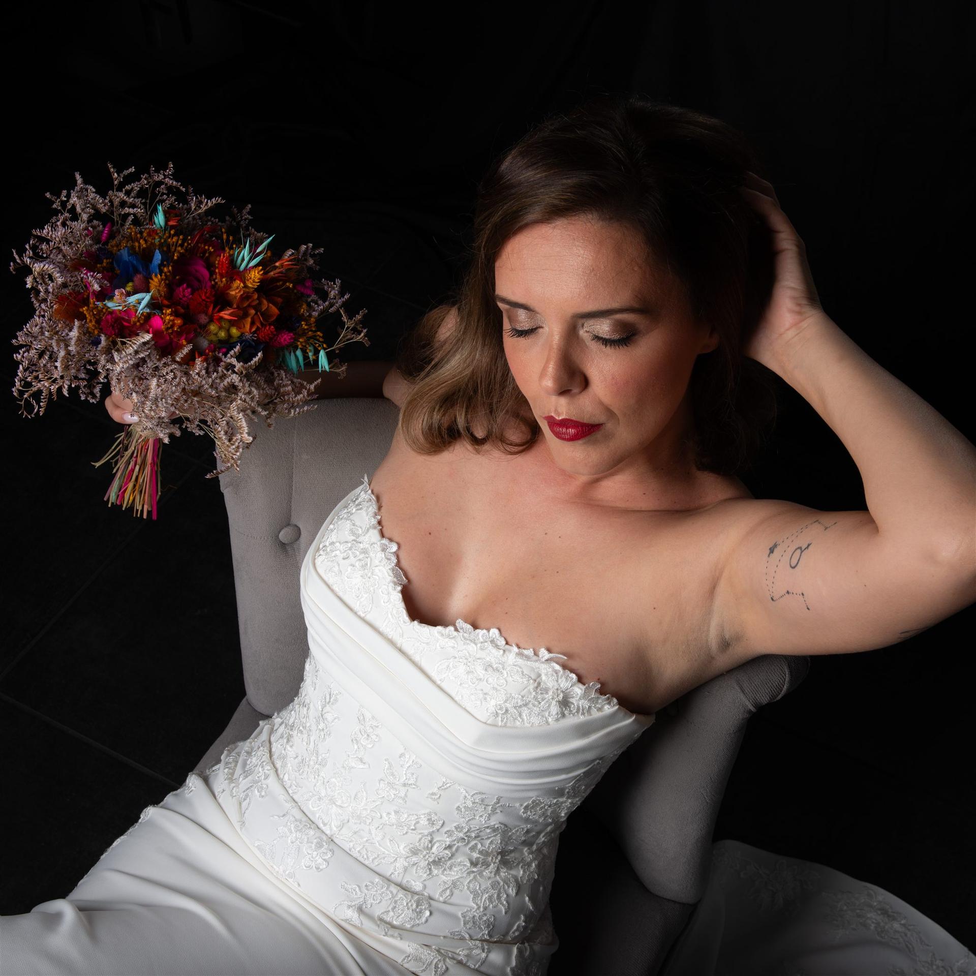 Portrait de la mariée dans un fauteuil gris sur fond noir, bouquet coloré, regard mélancolique — shooting mariage Perpignan