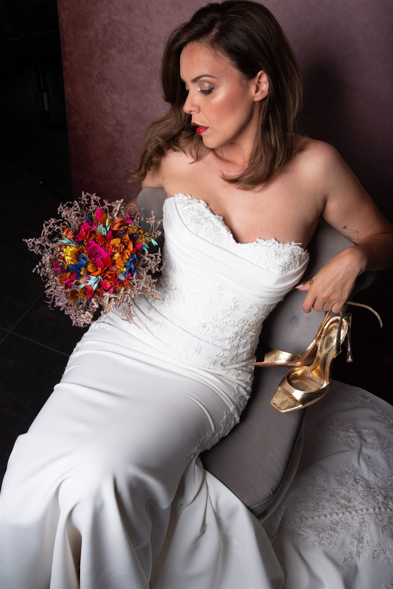 Portrait profil de la mariée dans un fauteuil, bouquet coloré, talon doré à la main — shooting inspiration mariage Perpignan
