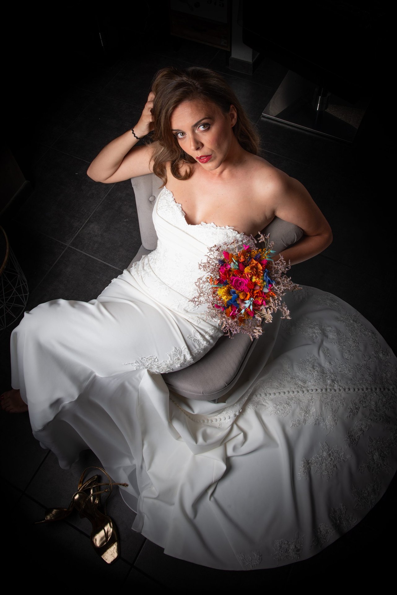Vue plongeante de la mariée dans un fauteuil gris, bouquet coloré, robe dentelle étalée — shooting inspiration mariage Perpignan